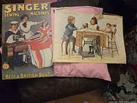 Photo showing two vintage Singer sewing machine signs: a large metal promotional sign and a smaller cardboard illustrated sign.