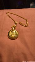 Gold-tone pocket watch closed showing embossed steam locomotive design on cover with attached chain laid on pink surface.
