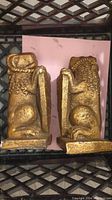 Pair of gold lion bookends shown standing upright on a black metal grid with a pink paper underneath.