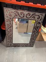 Full view of the rectangular mirror showing mosaic tile patterned border in shades of brown and beige placed on floor in storage area.