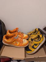 Photo of two pairs of Nike Dunk Low sneakers placed on shoeboxes against a white wall. The left pair is Magma Orange, showing signs of wear and moderate dirt on toe areas. The right pair is Goldenrod yellow with navy swooshes, showing moderate use and some discoloration from previous food stain.