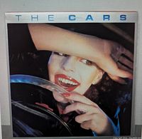 Front cover of The Cars vinyl album showing artwork of a person with hand partially covering face, smiling.