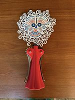 Top view of the Anna G. corkscrew showing the decorative white face with blue, orange, and red details and red dress body on a wooden surface.