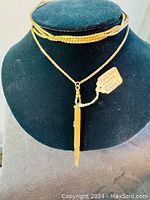 Gold plated chain draped on black mannequin, pencil pendant hanging at center with attached tag