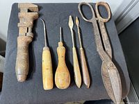 Top-down view of six hand tools arranged on black fabric, showing wrench, awls, spatulas, and scissors