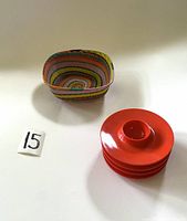 Photo showing four bright red/orange plastic egg cups stacked and the small multicolored woven basket nearby on a flat surface with a lot number card 15.