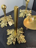 Two vertical ornate brass towel holders and one brass soap dish with all having leaf design bases, shown from diagonal overhead angle.
