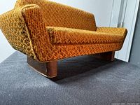 Angled side view showing textured golden-brown upholstery, right armrest, and wooden legs of the miniature sofa sample.