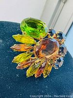 Close-up image of the vintage brooch showing large faceted green and amber glass stones with smaller amber and dark stones arranged in a prong setting