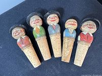 Front view of five cork wine stoppers with hand carved painted folk character tops.