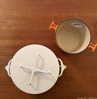 Top view of Dansk covered saucepan next to empty Le Creuset fondue pot on wooden surface.