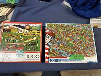 Photo showing two 1000-piece puzzles in original boxes: Charles Wysocki Buffalo and Where's Waldo themed puzzle.