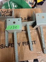 Three brand new metal deck anchors with attached rebar stakes shown on cardboard box.
