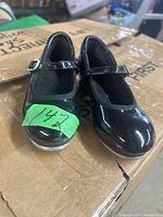 Front view of the pair of black patent finish girls tap shoes with ankle straps and buckles, placed on a cardboard box.