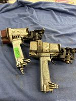 Two used pneumatic air nail guns lying on a blue fabric background, showing their sides and connection points. Both are visibly worn with dirt and some rust, intended for parts or repair.