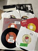 Photo showing several 45 RPM vinyl records, some in paper sleeves and others loose, with visible labels and barcode stickers.