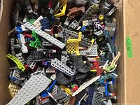 A box containing a large quantity of varied LEGO pieces including wheels, grey and colored bricks, mini-figure parts, and miscellaneous components in used condition.