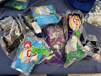 Photo showing 11 individually wrapped McDonald's kid's toys, all in original sealed plastic packaging with various McDonald's branding and numbered labels, placed on a blue surface.