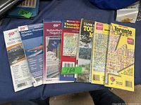 Photo of seven folded and booklet road maps spread out on a blue cloth surface. Shows maps from AAA and Toronto road maps with different covers and titles.