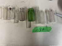 Photo showing 11 small glass bottles arranged in a row on a white surface, including one green bottle and one bottle with stopper.