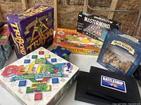 Photo showing boxes of several vintage board games including Tripbond, Mastermind, Blur, Operation, Brain Benders, and Battleship games arranged on a shelf.