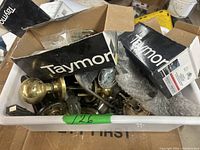Plastic bin filled with various used door hardware including brass door knobs, lock parts, and faceplates, with original Taymor boxes visible.
