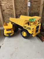 Yellow Tonka style dump truck and smaller yellow construction vehicle on concrete floor against wood wall background. The dump truck is large with a spacious dump bed, black wheels with yellow rims, and 'Fast Lane' text on the side.