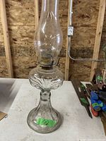 Photo of a vintage glass oil lamp with clear glass chimney and flared decorative base on a white table in a workshop setting.