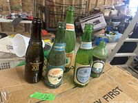 Photo showing seven vintage beer bottles in varied colors and labels placed on a cardboard box in a rustic setting.