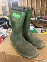 Pair of green Vulcan rubber boots, showing the left and right boot standing side by side on a cardboard surface in a workshop environment.
