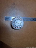 Baseball placed on brown surface next to a metal ruler measuring approximately 3 inches in diameter. Visible autographs include various names in black ink on white leather baseball with red stitching.
