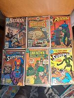 Top view of six comic books, each in protective plastic sleeves showing front covers including Batman, Hercules Unbound, Robin, Superman (two issues), and Unknown Soldier.