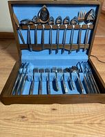 Full view of the flatware set displayed in a wood case with blue fabric lining, showing knives, forks, spoons, and serving utensils.