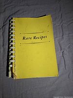 Yellow spiral-bound softcover cookbook titled 'Rare Recipes' showing use and stains on front cover.