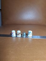 Front view of four thimbles with ruler for scale on leather background. Three ceramic with printed designs, one metal silver-colored.