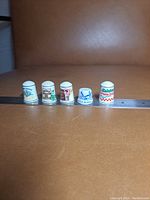 Front view of five collectible porcelain thimbles arranged in a row on a flat surface with an adjacent metal ruler showing size reference. Various colorful designs and logos on white thimble surfaces.