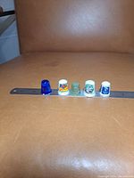 Five collectible thimbles arranged side by side on a brown surface with a ruler for scale measurement showing about 1 inch height each.