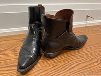 Pair of black leather cowboy boots showing pointed toes, boots angled with one standing upright and the other lying sideways on hardwood floor.