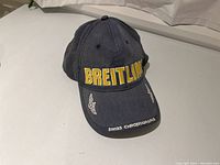 Dark blue Breitling baseball hat with yellow 'BREITLING' embroidery across front, white 'SWISS CHRONOGRAPHS' text on bill, and winged logos on sides of bill.