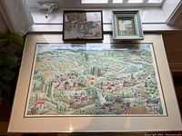 Photo showing a large, detailed framed village landscape print with vibrant greenery and cultivated fields, mounted under glass.