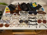 Wide view of 32 vintage eyeglasses and sunglasses spread on a table with their cases and cleaning accessories visible.