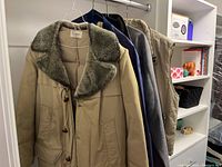 Photo showing four men's jackets on hangers, including a tan jacket with fur collar and button closure, beige sleeveless vest, gray zip-up jacket, and navy blue jacket.