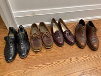 Four pairs of men's shoes lined up on wooden floor: black dress shoes with buckle, brown deck shoes, dark brown tassel loafers, and brown lace-up dress shoes.
