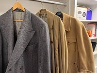 Three men's coats hanging on rack: gray herringbone Bill Griner, tan Aquascutum London, camel Cambridge.