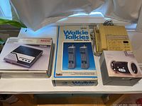 Photo showing vintage Sanyo answering system box and unit, AT&T walkie talkies original box, beige push-button telephone, and digital alarm clock.