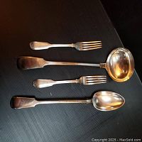 Four silver antique serving pieces including two forks, ladle, and serving spoon laid out on table.
