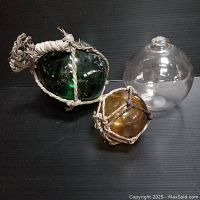Three glass fishing floats on black surface, one large green float with netting, one medium clear float, one small amber float with netting