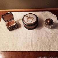 Three trinket boxes on cloth: metal Jennings Brothers box open showing satin lining, ceramic Toyo box with jockey on horse lid, small glass box with decorative metal lid.