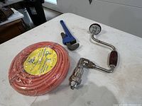 Four items on table: orange coiled extension cord, blue pipe wrench, chrome Millers Fall hand brace with bit and pipe wrench attachment