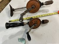 Two vintage Stanley woodworking hand drills with orange gear casing, black wooden handle and metal T-handle, placed on white table next to measuring tape.